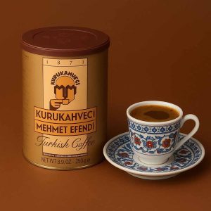 قوطی ۲۵۰ گرمی قهوه ترک مهمت افندی در کنار یک فنجان قهوه ترک روی پس‌زمینه قهوه‌ای.